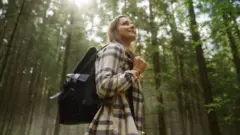 Kobieta spaceruje po lesie nordyckim. Ma na sobie jasną koszulę w kratę i czarny plecak