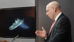 John Healey at a lectern with a picture of the Yantar behind