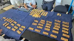 Centenas de moedas de ouro e dezenas de barras de ouro estão dispostas em pilhas sobre uma mesa coberta por um tecido azul-escuro. Dois policiais aparecem sentados à mesa, um deles usando um notebook.