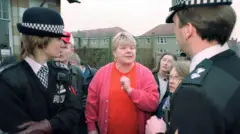 Mags Haney outside her home in the Raploch talking to two police officers. The photo from the mid 1990s shows Haney with short bleached blond hair and big earrings. She is wearing a pink cardigan and and orange t-shirt. A number of locals are standing around watching the scene