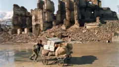 Jovem afegão empurra um carrinho carregado com sacos e caixas ao longo de uma rua de barro. Atrás dele, há uma construção destruída e entulho.