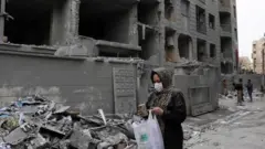 がれきの前を、マスクをした女性がスマートホンを見ながら歩いている。手には白いプラスチックバッグを持っている