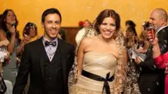 Jossette Rivera e seu marido Juan Fernando no dia do casamento dos dois. Ele veste terno preto e gravata borboleta, ela veste um vestido de noiva bege tomara-que-caia, com um laço preto na cintura e véu bordado. Os convidados sopram bolinhas de sabão à volta do casal