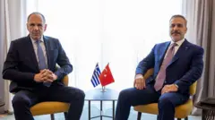 Türk ve Yunan bakanlar yan yana otruyor. İki ülkenin küçük bayrakları aralarındaki yuvarlak bir masada duruyor.