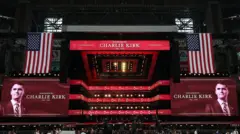 Um palco de estádio coberto, com grandes bandeiras americanas de cada lado e duas projeções do rosto e nome de Charlie Kirk com os dizeres: "Em memória de Charlie Kirk, 1993 - 2025".