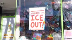 Une pancarte rouge indiquant "Les voisins disent : ICE OUT !" est accrochée à un verre dans une boutique de souvenirs.