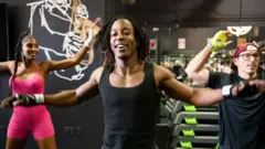 Três pessoas participam de uma aula de dança fitness em uma academia, realizando movimentos sincronizados com os braços abertos. O instrutor, ao centro, sorri enquanto lidera a atividade, acompanhado por dois alunos ao fundo, em um ambiente com equipamentos de musculação.