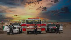Tres funcionarios uniformados están parados frente a tres vehículos oficiales que incluyen un auto de patrulla, un camión de bomeros y una camioneta de vilancia. Un dron flota sobre ellos y en el fondo se ve el paisaje desértico en la luz del atardecer