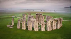Resim Stonehenge'i gösteriyor. Arkasında antik anıtın tam dairelerinin ön planda olduğu sisli bir gökyüzü var. Dış dairenin bir parçasını oluşturan, her biri üstte başka bir büyük taş bulunan iki büyük dik taştan oluşan dört set. İç alanın daha büyük taşları dış çemberinkilerin üzerinde görülebilir. Yeşil çim anıtı her taraftan çevreliyor.