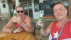 Longworth (esquerda) e Alfonso (direita), sorriem para a câmera. Eles seguram garrafas verdes de cerveja Stella Artois e vestem camisetas vermelhas com desenhos azuis e brancos. Eles estão sentados no que parece ser um bar ou restaurante em ambiente externo, observando a paisagem.