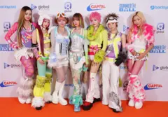 Fotografia colorida mostra os integrantes do XG posando em frente a um pôster que promove o Summertime Ball. Eles usam roupas de cores vibrantes, além de uma variedade de chapéus e botas com acabamento em pele.