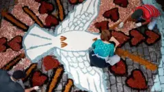 Foto de cima mostra uma mulher no chão fazendo um tapete de Corpus Christi em uma rua.