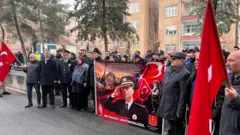 Yaklaşık 40-50 kişi Gaffar Okkan'ın posteri ve Türk bayraklarıyla anmak için duruyor. Aralarında polisler ve siviller görülüyor