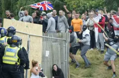 Uma mulher e um homem se abrigam enquanto objetos são lançados durante um protesto anti-imigraçãom betfrente ao Holiday Inn Expressm betRotherham, South Yorkshire