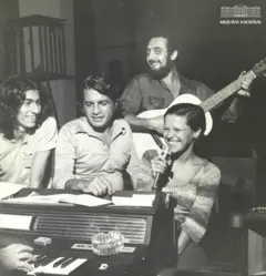 Elis Regina sorrindo ao lado de homens, a maioria sentados; é possível ver alguém ao piano e homem em pé com violão