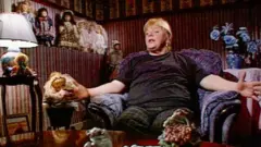 Imagem de Haney em sua casa, tirada de um documentário do início dos anos 2000. Ela está sentada em uma poltrona roxa, cercada por bonecas