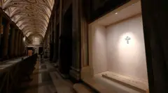 Vista do túmulo do Papa Francisco em um salão da Basílica de Santa Maria Maggiore, com teto abobadado e pilares de mármore. Um holofote solitário ilumina um crucifixo acima da lápide.