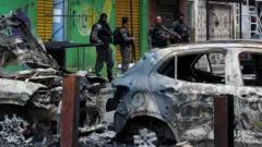 Policias fortemente armados observam carcaças de carros encendiados