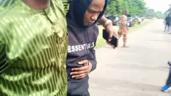 Kayode Adeniyi wọ aṣọ dudu, eeyan kan wọ aṣọ alawọ ewe fi ọwọ kọ lọ́run