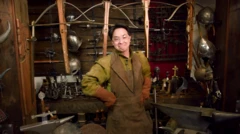 Ash em uma oficina de espadas. Atrás deles há vários arcos e flechas, espadas e outras armas.