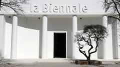 Bela zgrada sa natpisom „la Biennale“ i crnim drvetom ispred nje