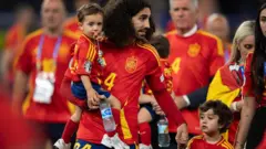 Cucurella com a camisa da Espanha de mãos dadas com o filho Mateo e segurando o outro filhi no colo