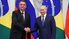 Bolsonaro e Putin sorrindo e apertando as mãos, em frente a bandeiras do Brasil e Rússia