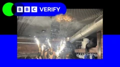 BBC Verify 로고가 있는 브랜드 이미지. 푸른색 테두리가 둘러진 사진 속에는 스위스 스키 리조트의 바에서 사람들이 스파클라가 부착된 샴페인 병을 들고 있는 모습이 담겨 있다. 이는 화재가 발생하기 직전의 장면으로, 스파클라 바로 위 폼이 설치된 천장에는 작은 주황색 불꽃 자국이 보인다.