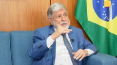 Celso Amorim usando terno azul claro com uma bandeira do Brasil ao fundo