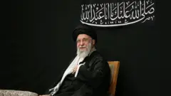 Ayatollah Ali Khamenei is pictured seated against a black backdrop, dressed in black robes and looking to the left of the camera. 