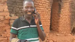 Umugabo umwenyura urimo kuvugira kuri telefone ari imbere y'inzu y'amatafari