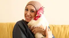 تجلس فوزية على أريكة صفراء، وهي تحتضن ديكها الأبيض
