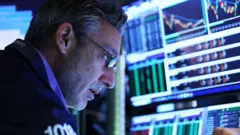 Homem olha atento para telas de computador, com gráficos e números sobre as oscilações das bolsas de valores.