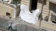 Вход в здание, поврежденный при взрыве, прикрытый белым полотном, перед зданием идет полицейский.