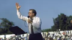 Jimmy Swaggart hold microphone dey preach to one large crowd outside during di 1980s.