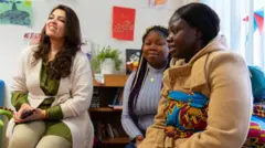 Três mulheres sentadas em uma sala, sorrindo. As paredes atrás delas estão decoradas com desenhos coloridos de crianças