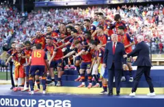 رودري، لاعب المنتخب الإسباني رقم 16، يستعد لرفع كأس العالم لكرة القدم، بينما يوجه رئيس الفيفا جياني إنفانتينو الرئيس الأمريكي دونالد ترامب، للخروج من أمام المنتخب الإسباني المحتفل بفوزه.
يرتدي ترامنب وانفانتينو بدلات رسمية، بينما يرتدي لاعبو المنتخب الإسباني لباس منتخب بلادهم الرياضي بالألوان الأحمر والكحلي والأصفر، يحتفلون وخلفهم الجماهير.
