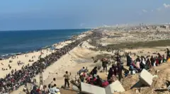 آلاف النازحين يعودون إلى شمال غزة بعد وقف إطلاق النار، في الصورة الملتقة نهاراً الناس بأعداد كبيرة يقفون قرب الشاطئ، ويرفع أحدهم العلم الفلسطيني على برج يبدو أنه للاتصالات أو الكهرباء