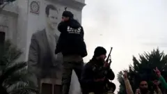 مقاتلون مسلحون يقفون أمام لافتة عليها صورة الرئيس السوري بشار الأسد في حماة.