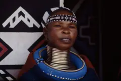 Imagem de uma mulher com anéis dourados empilhados ao redor do pescoço, da base até o queixo. Ela está usando uma faixa de cabeça com contas.