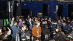 Fila para entrar no bar em um ambiente escuro. A maioria dos clientes é formada por homens jovens