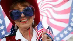 Una mujer con una sombrilla con los colores de la bandera de Estados Unidos, vestida con un sombrero estilo vaquero rojo, chaleco de lentejuelas rojo y azul, camisa blanca y unos lentes azules y rojos. Lleva en su mano dos pequeñas banderas de Estados Unidos. 