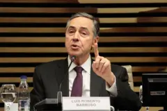 Barroso gesticulando enquanto fala, sentado, em salão; à sua frente, placa com seu nome