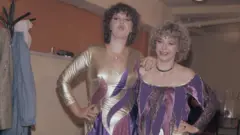 Duas concorrentes do Intervision de 1977 posam para a câmera nos bastidores. A mais alta, com cabelos castanhos, franze os lábios. A mais baixa, com cabelos loiros, sorri. Ambas vestem trajes combinando nas cores dourado e roxo. Ao fundo, vê-se um cabide com casacos.