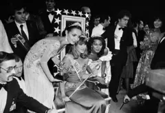 Mulher sentada em uma cadeira com uma coroa. Há várias pessoas ao redor