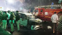 インド警察の隊員が自動車爆発現場を捜索している。白い焼け焦げた車と、背景に赤い消防車が見える