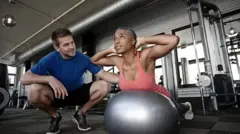 Uma mulher em cima de uma bola grande de pilates e um homem ao lado dela instruindo