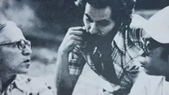 Foto em preto e branco mostra três homens conversando, com olhares compenetrados