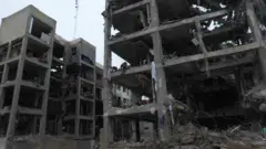 Dois prédios destruídos, com quatro ou cinco andares, com janelas e paredes estouradas. Algumas colunas e lajes de concreto ainda permanecem. Há alguns pedaços de tecido pendurados na estrutura e alguns metais retorcidos no chão.