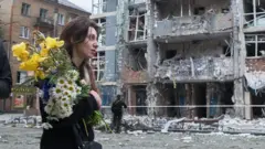 امرأة تحمل باقة من الزهور أمام مبنى سكني شاهق الارتفاع في منطقة متضررة بشدة جراء غارة روسية بطائرة بدون طيار في كييف في 25 مايو/ أيار 2025